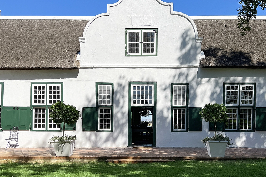 Perfect Hideaways property for sale: A Cape Dutch homestead with green shuttered windows, featuring a terracotta stone patio outside the main entrance, and a manicured lawn in front of the house. Hopehill Manor Boutique Guesthouse, Stellenbosch