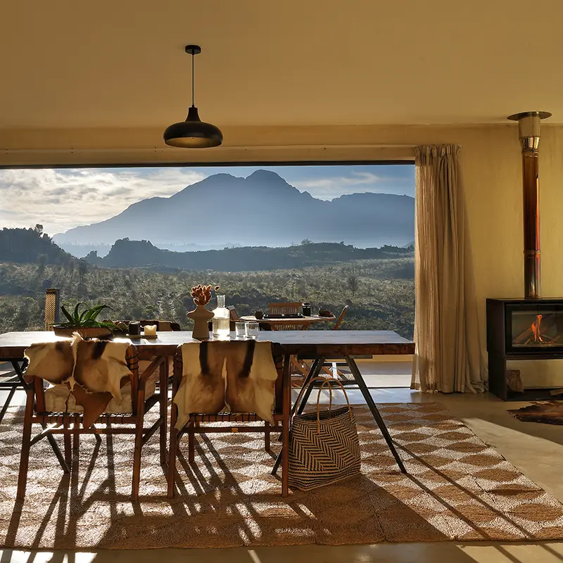 Perfect Hideaways property for sale: Interior dining area with a wooden table, chairs draped in fur, and a large window revealing a mountain view. A fireplace glows gently. Oomblik Cabin, Mount Bain Private Nature Reserve.