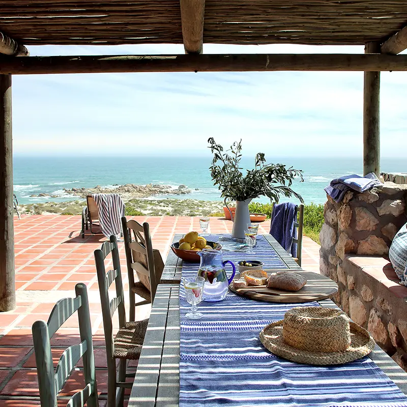 Perfect Hideaways property for sale: Outdoor dining scene on a patio with ocean view, featuring a striped tablecloth, bread, fruit, a sunhat, and relaxed wooden chairs under a wooden pergola. Whale Cottage in Paternoster.