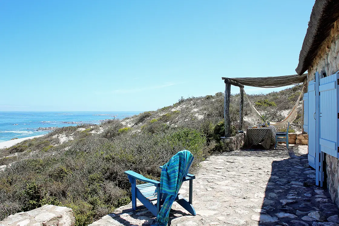 Perfect Hideaways property for sale: Stone patio overlooking the ocean, with a blue chair draped in a towel, table with chairs, a hammock, and rustic stone cottage. Artemon Cottage, Paternoster.
