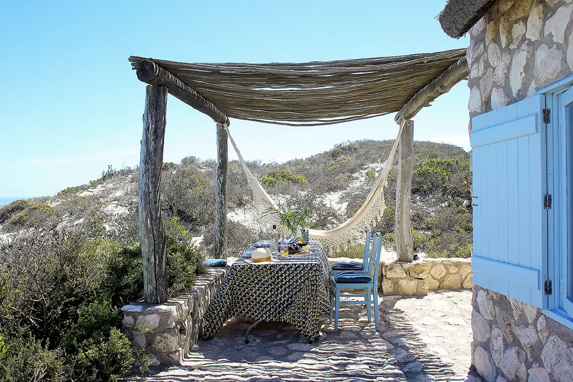 Perfect Hideaways property for sale: Outdoor dining scene with a rustic wooden pergola and hammock, under a clear blue sky. A table with a patterned cloth, blue chairs, and a hat is surrounded by sandy dunes and scrubby vegetation. Artemon Cottage, Paternoster.