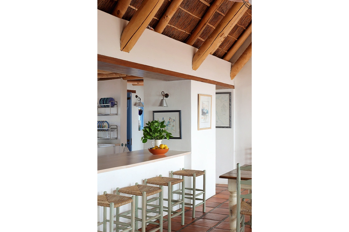 Perfect Hideaways property for sale: Rustic kitchen with a natural wood and thatch ceiling, wicker stools, and terracotta floor. A fruit bowl and plant on the counter add a fresh touch. Whale Cottage in Paternoster.