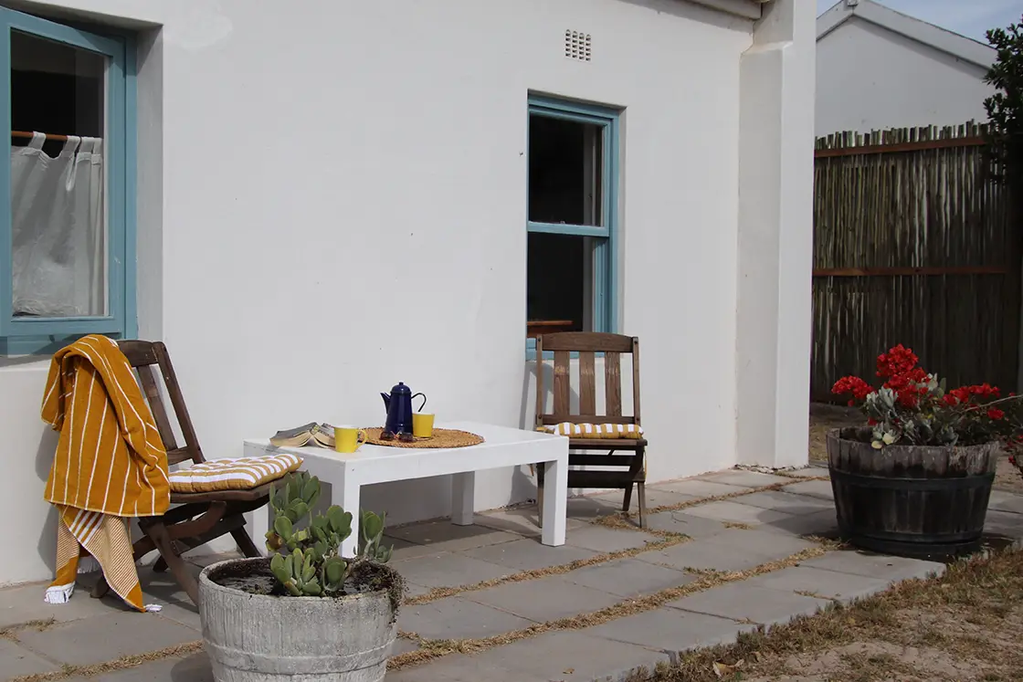 Perfect Hideaways property for sale: Outdoor patio scene with two wooden chairs, a white table, yellow-striped cushions, and an orange blanket. Potted plants add a cozy touch. The Cottage in Churchhaven.
