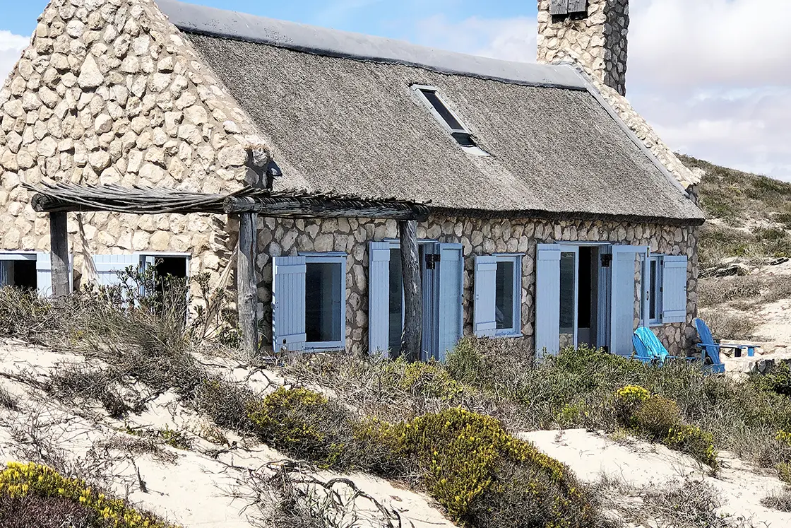 Perfect Hideaways property for sale: Stone cottage with blue shutters and a thatched roof nestled among sandy dunes and sparse vegetation, evoking a rustic seaside charm. Artemon Cottage, Paternoster.