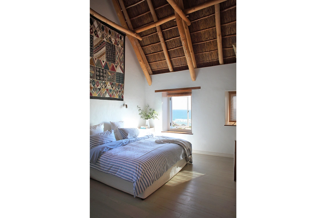 Perfect Hideaways property for sale: Cozy bedroom with a striped bed, white walls, and a wooden ceiling. A window offers an ocean view, with a vibrant patterned tapestry on the wall. Whale Cottage in Paternoster.
