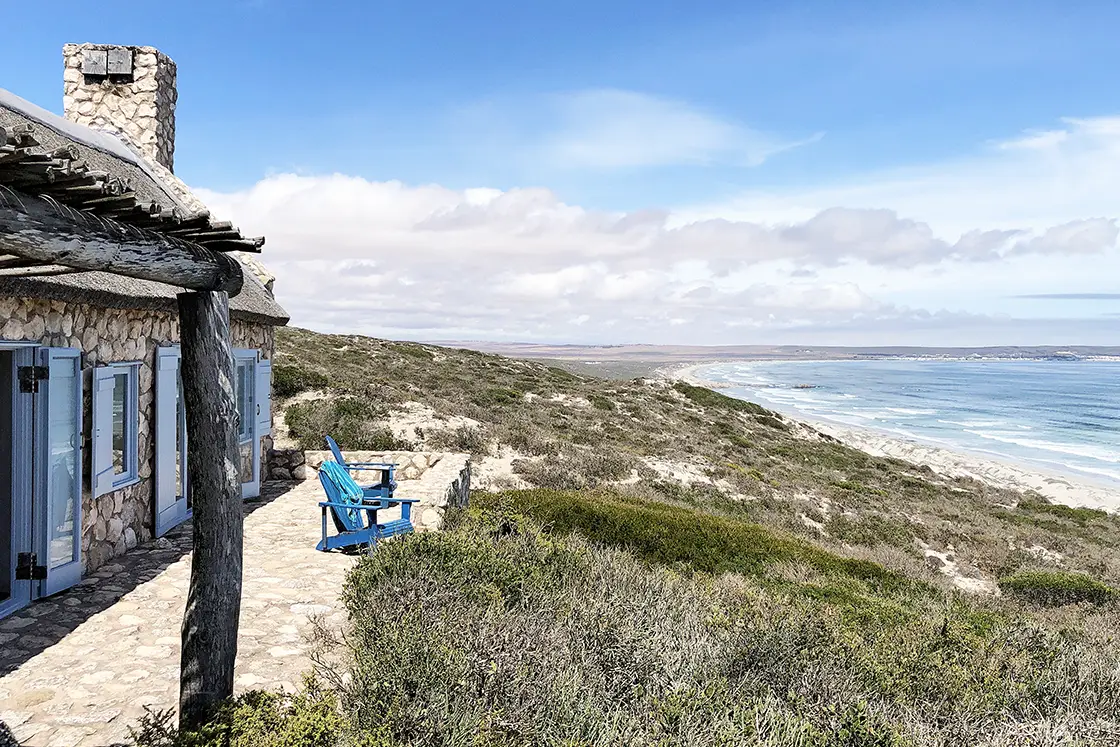 Perfect Hideaways property for sale: Stone cottage with blue shutters on a hill overlooking the ocean. A blue chair on the patio faces the waves. Artemon Cottage, Paternoster.
