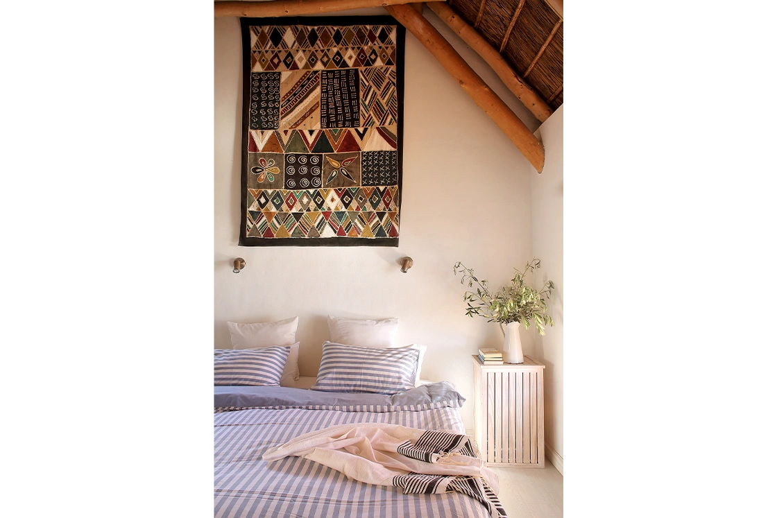 Perfect Hideaways property for sale: Cozy bedroom with striped bedding, African-patterned wall tapestry, and rustic wooden ceiling beams. A nightstand holds a vase with green branches. Whale Cottage in Paternoster.