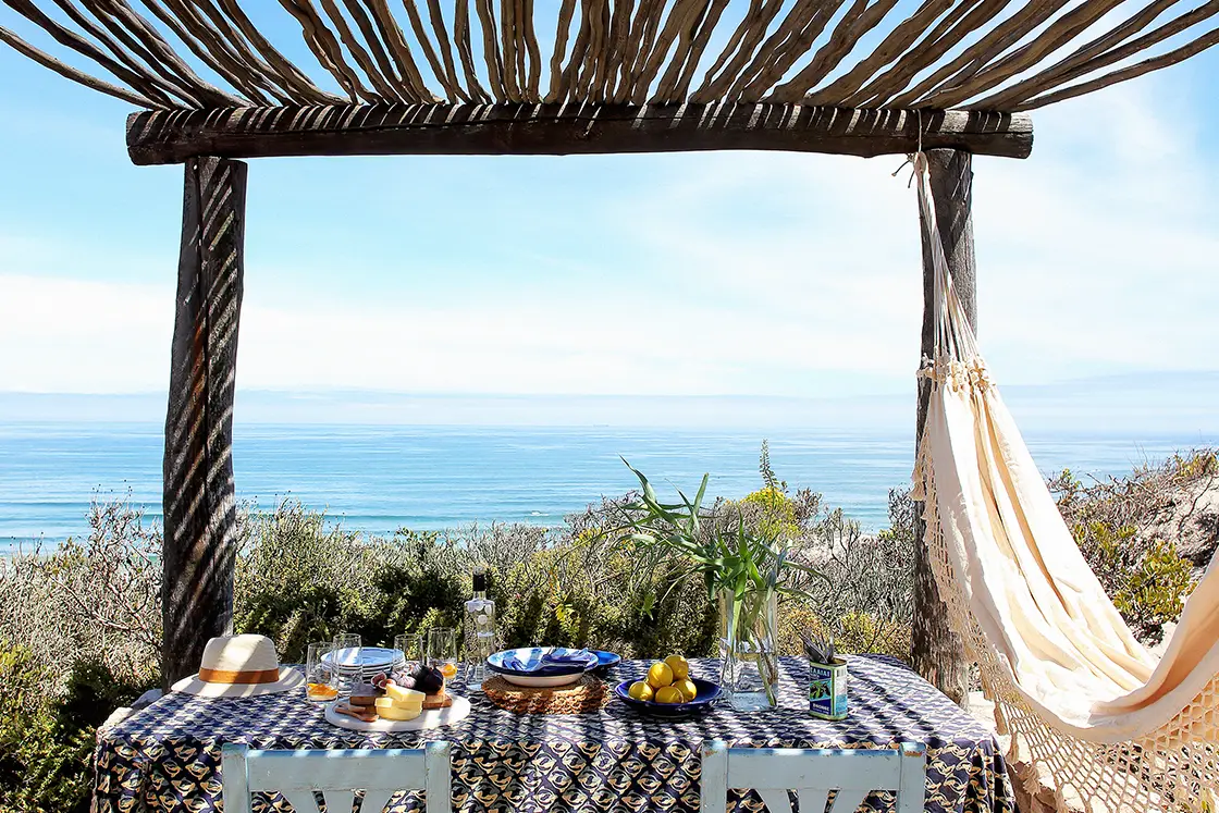 Perfect Hideaways property for sale: Cozy outdoor dining setup under a wooden pergola overlooking the ocean. A table with a patterned cloth holds plates, drinks, fruits, and a vase. A hammock swings nearby. Artemon Cottage, Paternoster.
