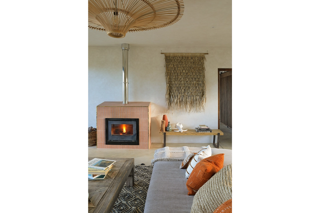 Perfect Hideaways real estate property: Cozy living room with a lit fireplace, textured wall hanging, wooden furniture, and orange cushions. Warm, inviting atmosphere with rustic decor. Wolwehoek Private Nature Reserve, Montagu.