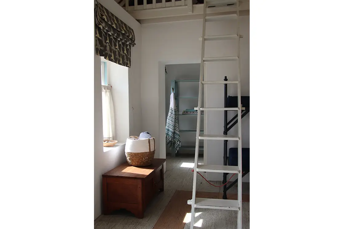 Perfect Hideaways property for sale: Loft with white ladder, rustic wooden chest, and a basket under a window with patterned curtains. The room is airy with light streaming in. The Cottage in Churchhaven.
