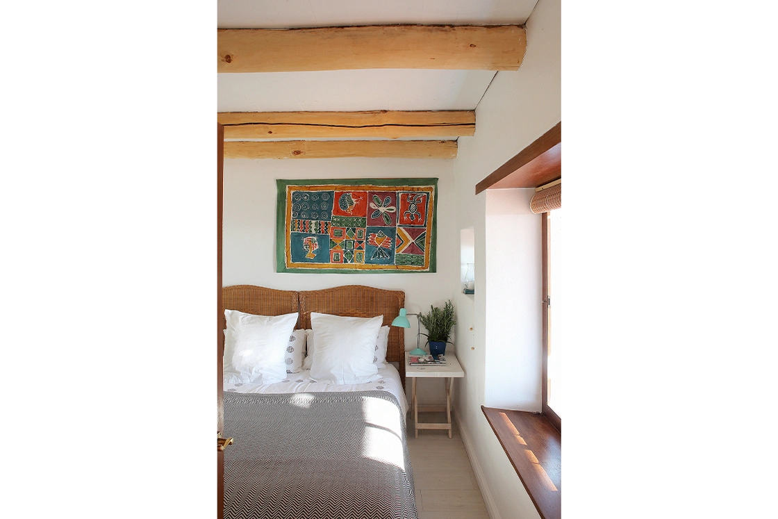 Perfect Hideaways property for sale: Cozy bedroom with a wicker headboard, white pillows, colorful wall tapestry, and wooden beams. Sunlight streams through a window, creating a warm tone. Whale Cottage in Paternoster.