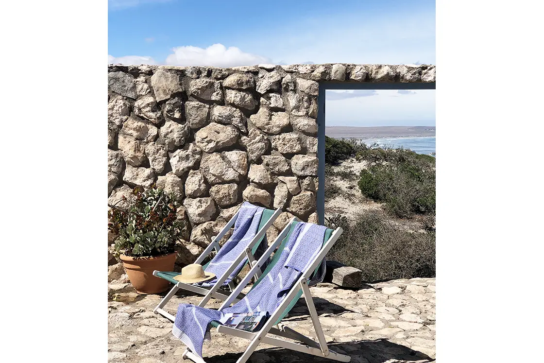Perfect Hideaways property for sale: Two deck chairs with towels and a hat, set on a stone patio by a rustic wall. Beyond, a serene coastal landscape with blue skies and distant sea. Artemon Cottage, Paternoster.