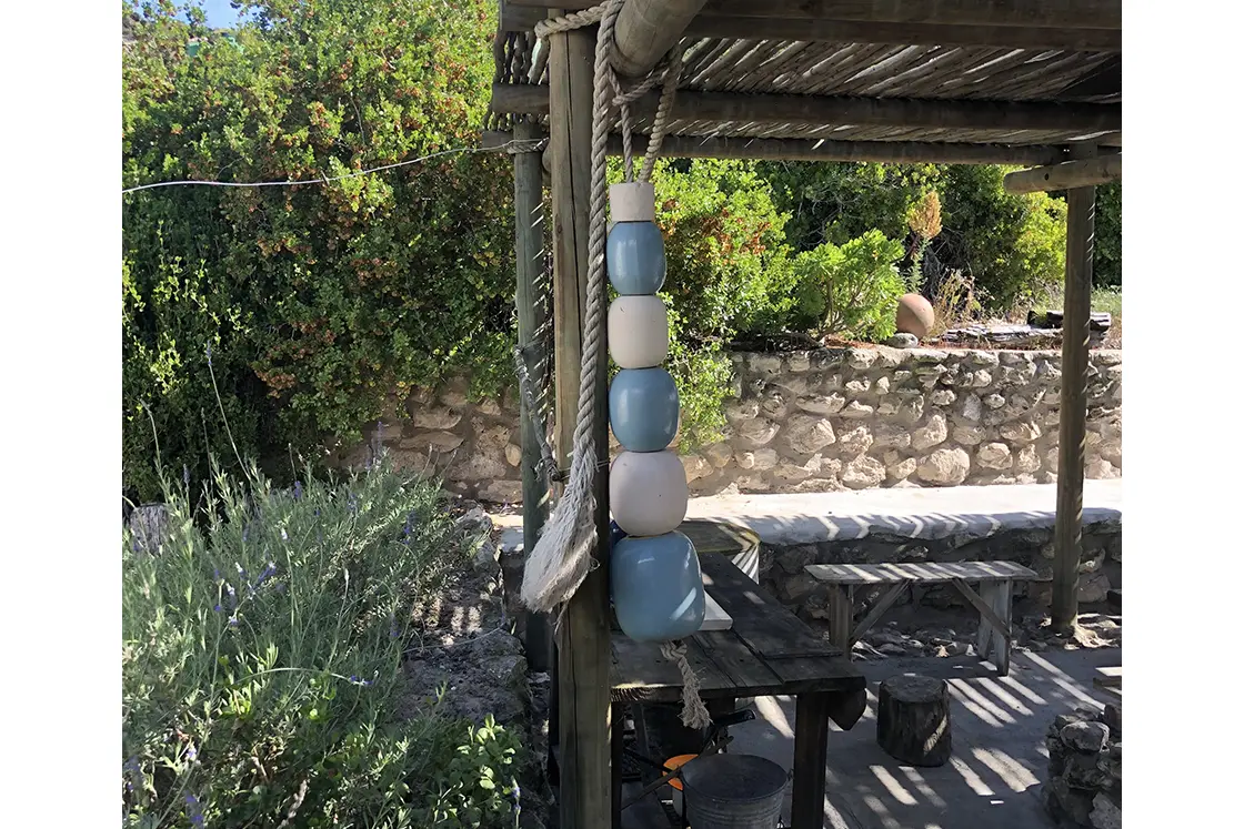 Perfect Hideaways property for sale: A rustic pergola with colorful stacked buoys hanging from thick ropes. Stone wall and vibrant green foliage in the background, creating a serene, coastal ambiance. The Cottage in Churchhaven.