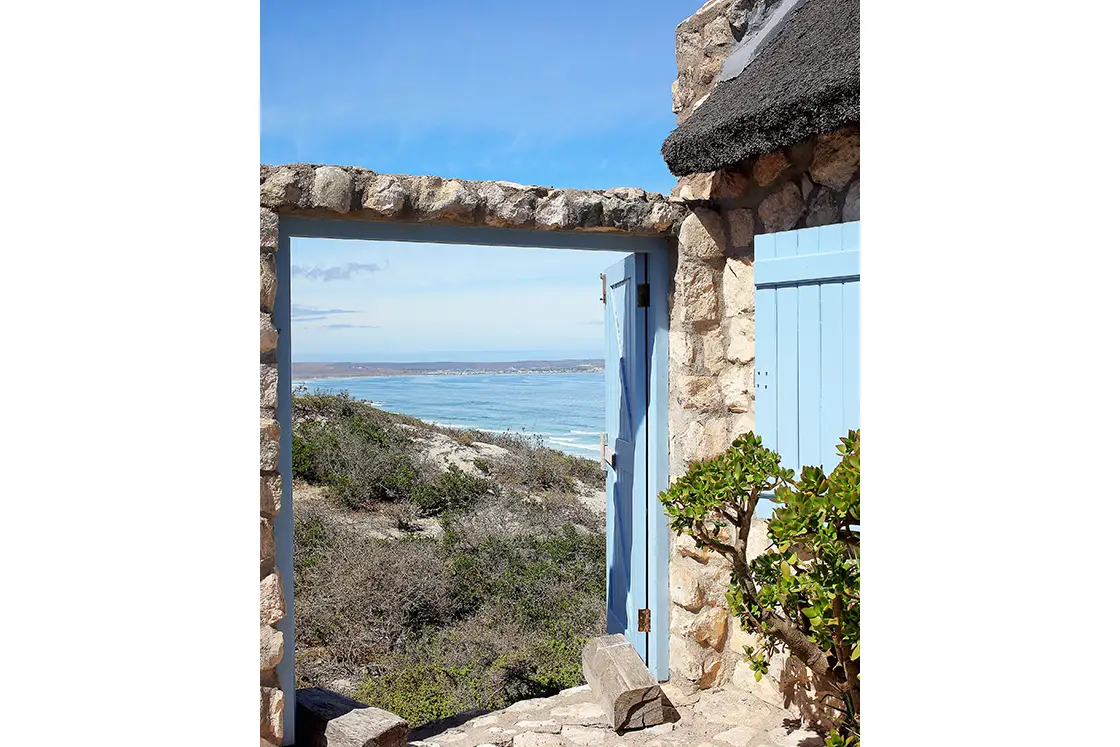 Perfect Hideaways property for sale: Stone doorway with a light blue wooden door open to a scenic ocean view. A bush grows nearby. Artemon Cottage, Paternoster.