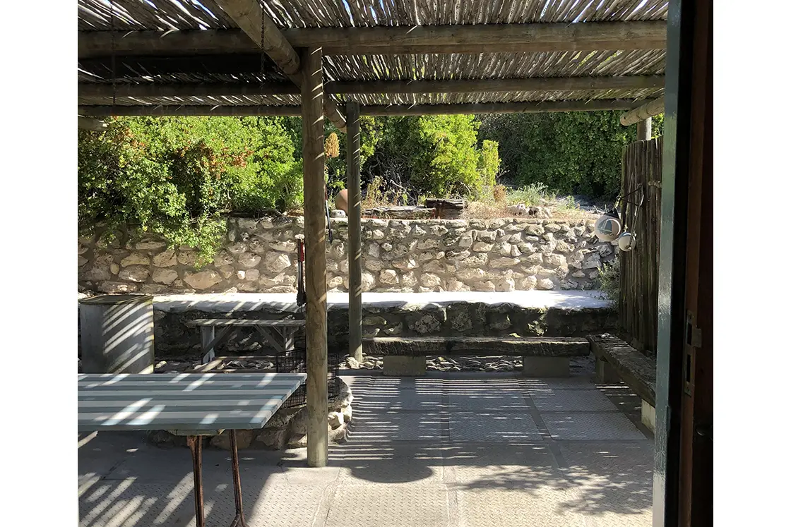 Perfect Hideaways property for sale: Shaded outdoor patio with wooden pergola casting striped shadows. Stone wall in background, lush greenery, and hanging cups create a serene, rustic vibe. The Cottage in Churchhaven.