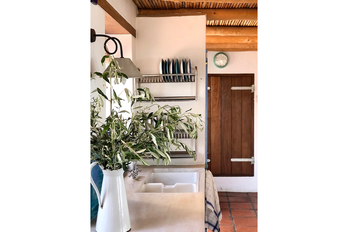 Perfect Hideaways property for sale: Cozy rustic kitchen scene with plants in a white jug by a wooden counter, sink, and stacked plates on a rack. A wooden door and clock add charm to the space. Whale Cottage in Paternoster.