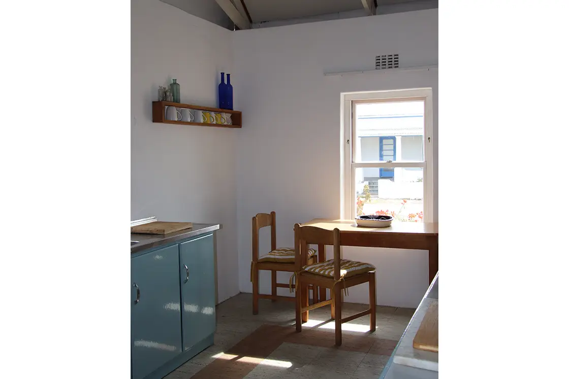 Perfect Hideaways property for sale: Cozy kitchen with natural light. Wooden table and chairs by a window, blue cabinet, floating shelf with mugs, glass bottles, creating a serene ambiance. The Cottage in Churchhaven.