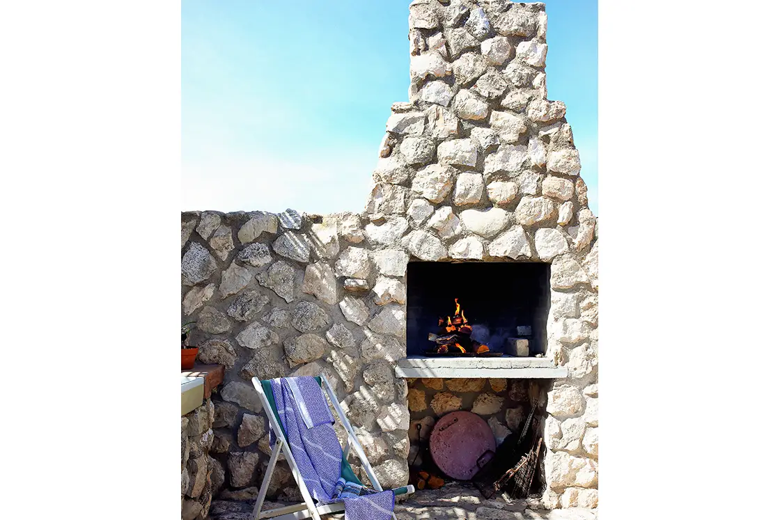 Perfect Hideaways property for sale: Stone outdoor fireplace with logs burning beneath a clear blue sky. A folded deck chair with a blue towel rests nearby. Artemon Cottage, Paternoster.