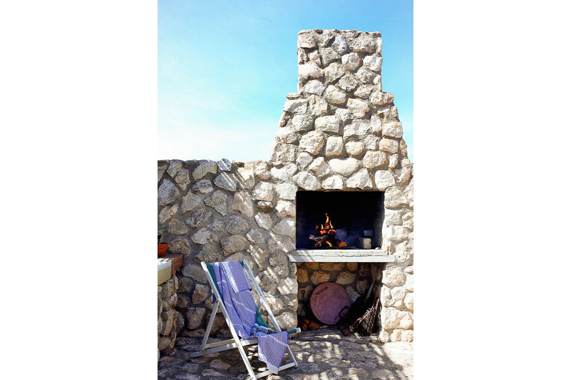 Perfect Hideaways property for sale: Stone outdoor fireplace with logs burning beneath a clear blue sky. A folded deck chair with a blue towel rests nearby. Artemon Cottage, Paternoster.