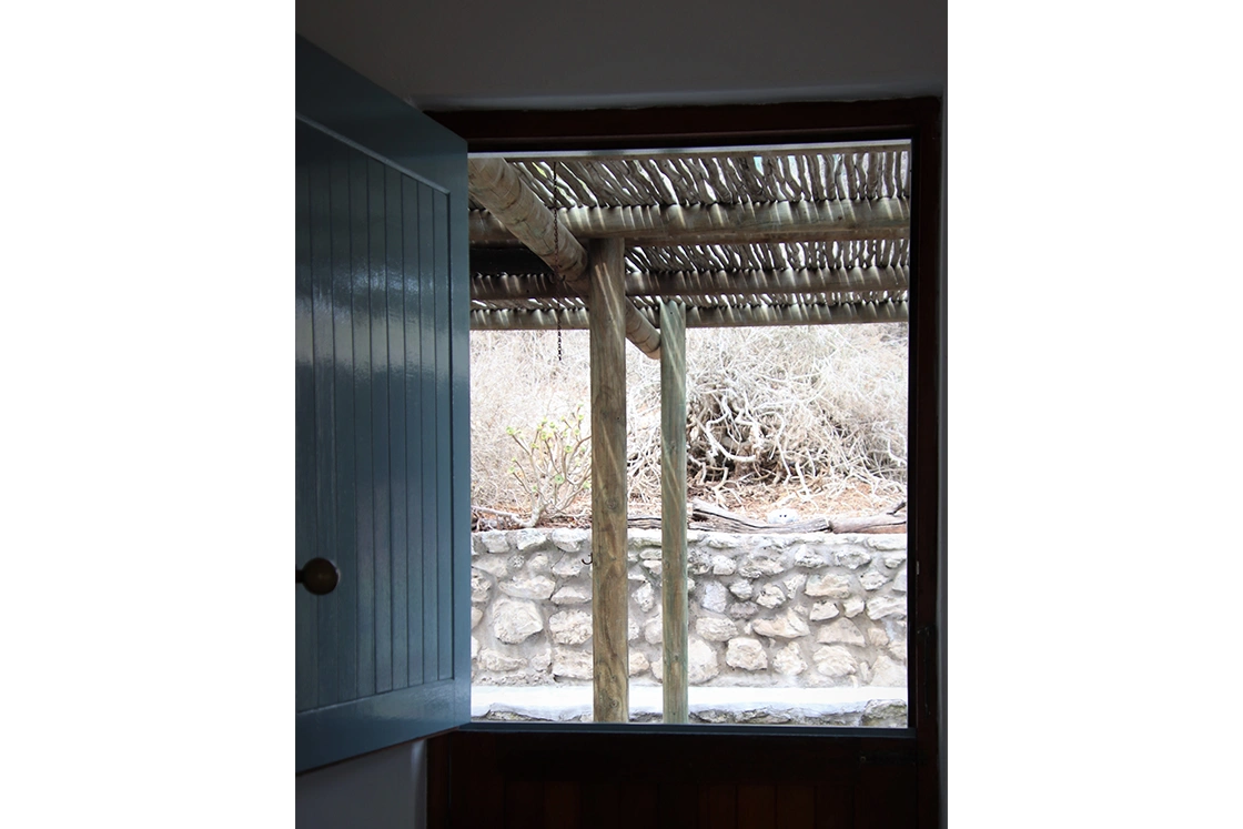 Perfect Hideaways property for sale: View from an open blue door shows a rustic outdoor scene with wooden poles supporting a pergola, a stone wall, and dry, tangled bushes beyond. Tranquil setting. The Cottage in Churchhaven.