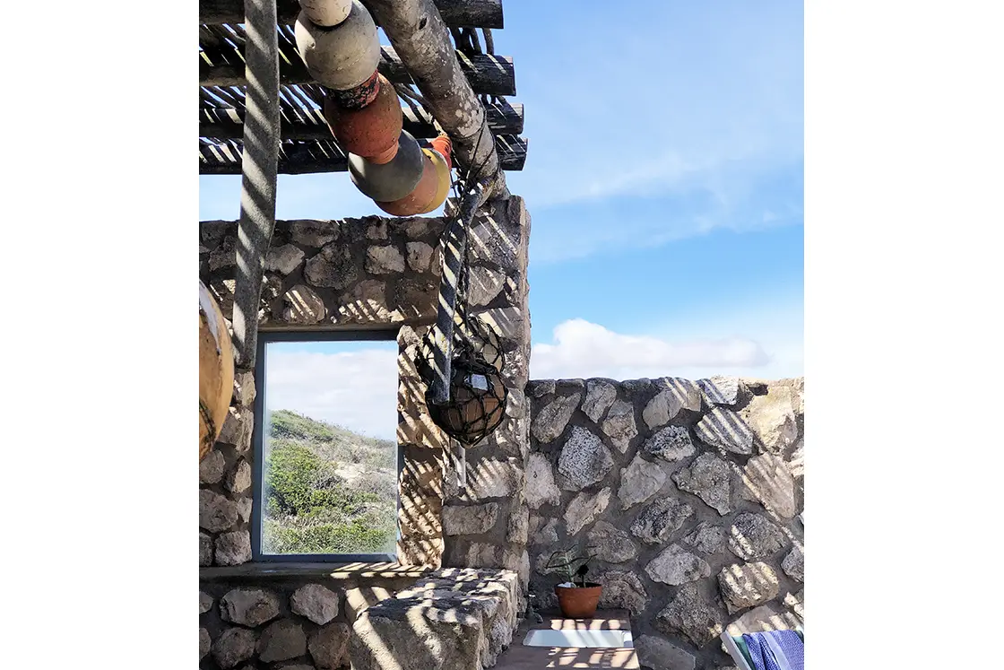 Perfect Hideaways property for sale: Stone structure with a wooden pergola casting striped shadows. Colorful buoys hang from ropes. A window offers a view of greenery under a clear blue sky. Artemon Cottage, Paternoster.