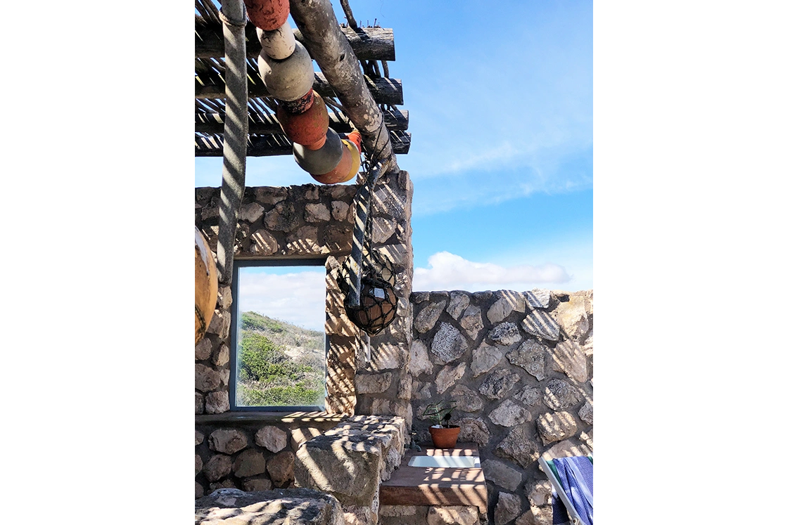 Perfect Hideaways property for sale: Stone structure with a wooden pergola casting striped shadows. Colorful buoys hang from ropes. A window offers a view of greenery under a clear blue sky. Artemon Cottage, Paternoster.