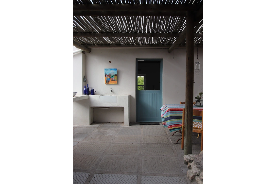 Perfect Hideaways property for sale: Outdoor patio with rustic wooden ceiling. Features a blue door, wall art, white sink, and a table covered with a colorful striped tablecloth. Cozy ambiance. The Cottage in Churchhaven.