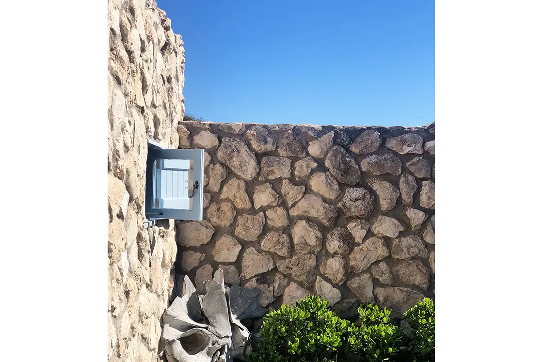 Perfect Hideaways property for sale: Stone wall in bright sunlight with a small open window, set against a clear blue sky. Green bushes and gray cement pieces at the base. Artemon Cottage, Paternoster.