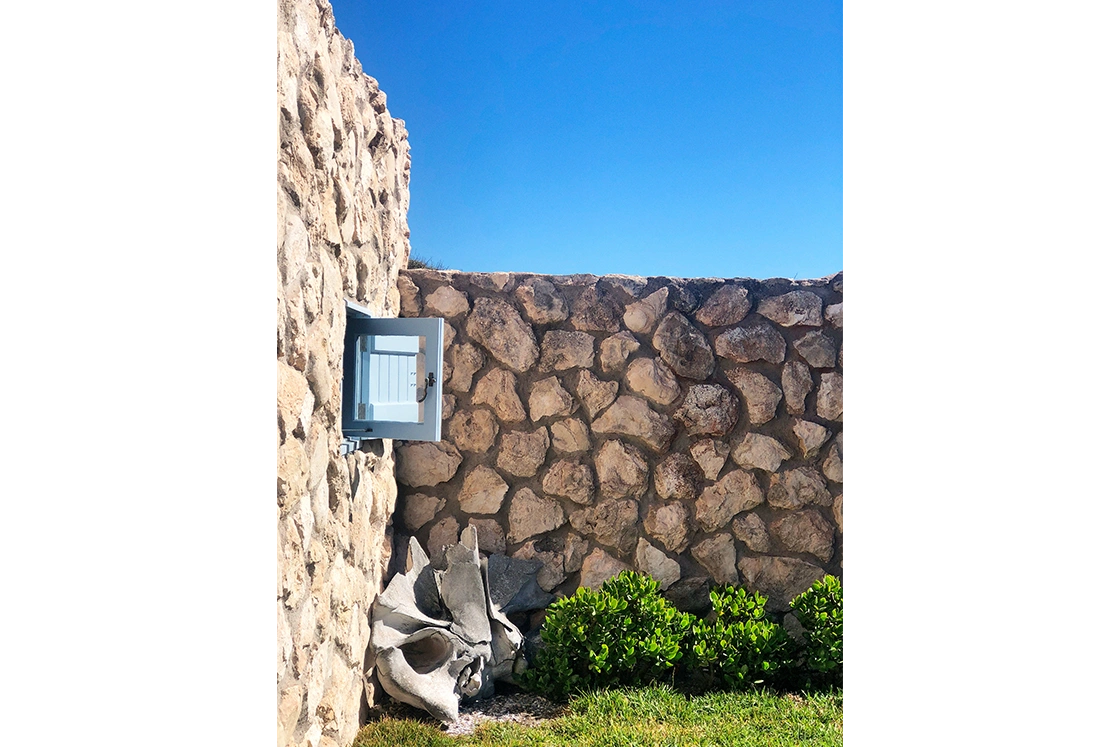 Perfect Hideaways property for sale: Stone wall in bright sunlight with a small open window, set against a clear blue sky. Green bushes and gray cement pieces at the base. Artemon Cottage, Paternoster.