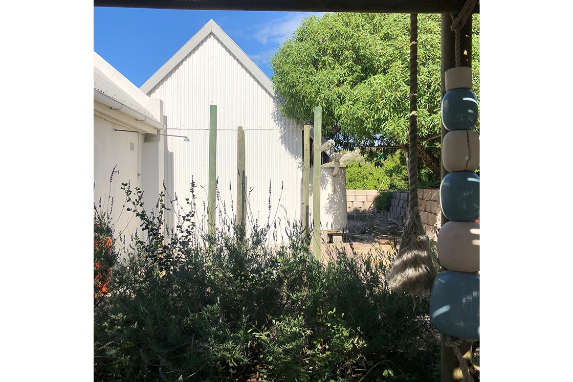 Perfect Hideaways property for sale: White building with a gabled roof stands in a sunny garden. Green shrubs and a tree create a tranquil ambiance. Rope with blue buoys hangs in the foreground. The Cottage in Churchhaven.