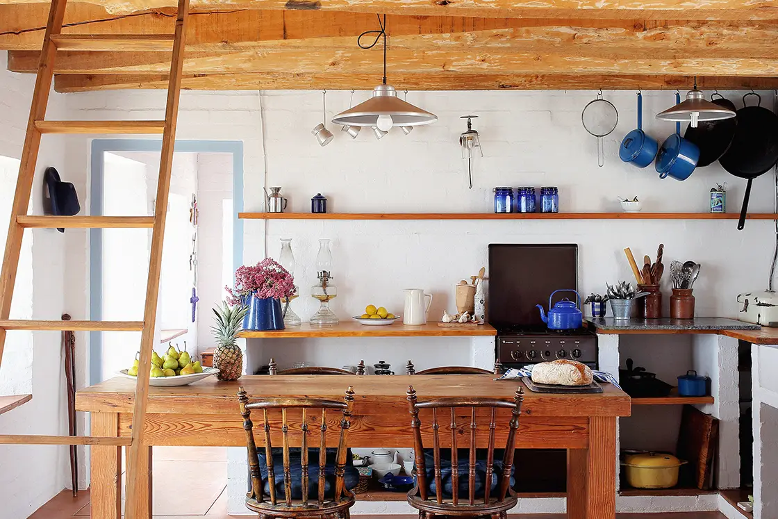 Perfect Hideaways property for sale: Rustic kitchen with wooden beams and shelves, featuring blue pots, hanging pans, and a table with flowers, fruit, and bread. Artemon Cottage, Paternoster.