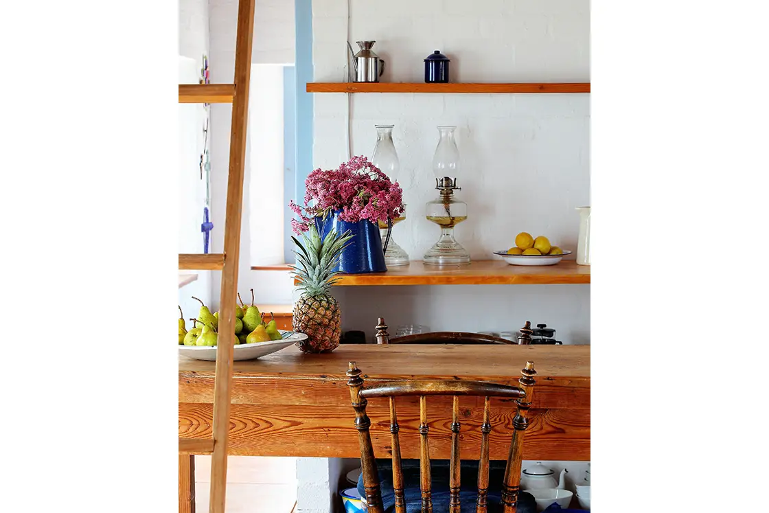 Perfect Hideaways property for sale: Cozy kitchen with wooden shelves displaying a blue vase of pink flowers, oil lamps, and a bowl of lemons. A table holds pears and a pineapple. Artemon Cottage, Paternoster.