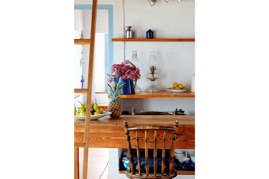 Perfect Hideaways property for sale: Cozy kitchen with wooden shelves displaying a blue vase of pink flowers, oil lamps, and a bowl of lemons. A table holds pears and a pineapple. Artemon Cottage, Paternoster.
