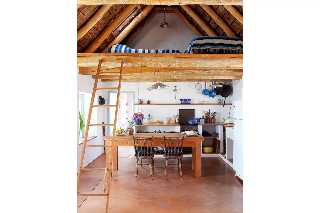 Perfect Hideaways property for sale: Cozy loft kitchen with rustic wooden beams and a ladder leading to a bed alcove. Wooden dining table, chairs, and kitchen shelves. Artemon Cottage, Paternoster.