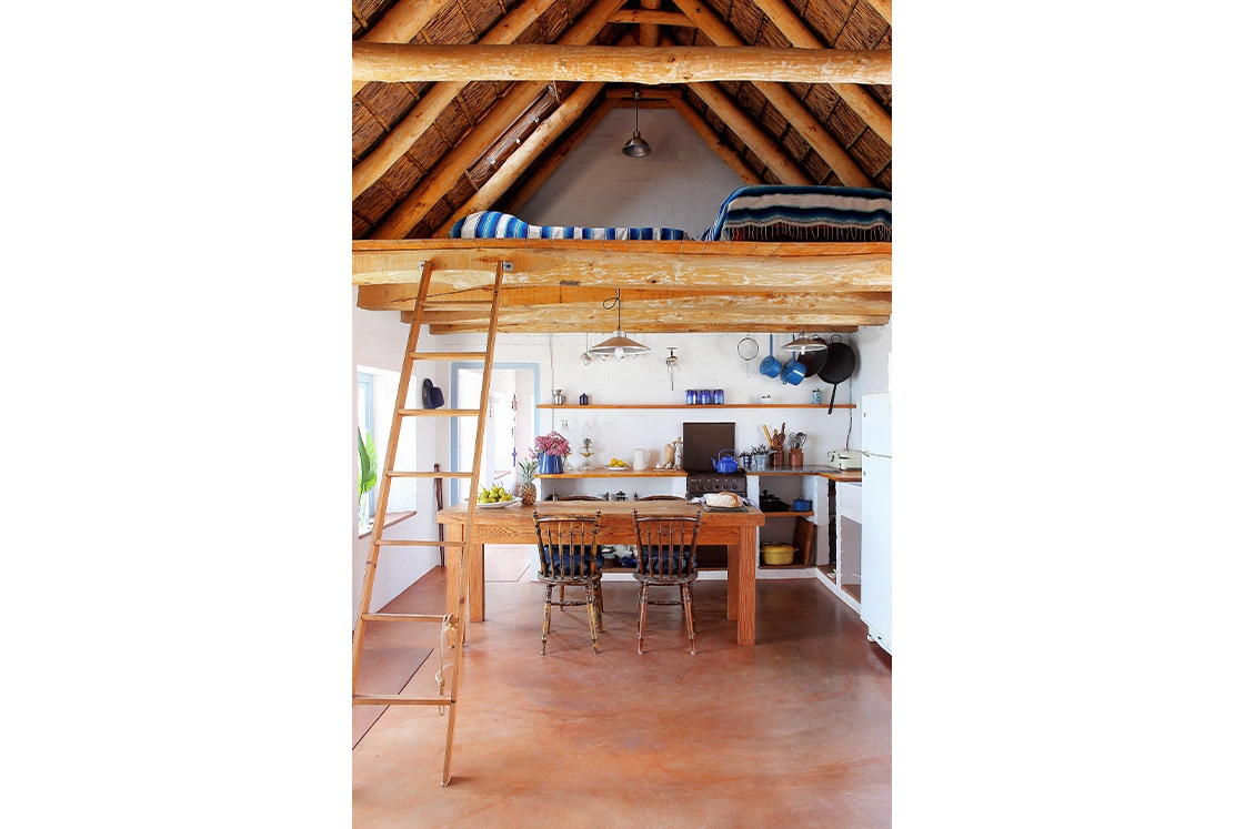 Perfect Hideaways property for sale: Cozy loft kitchen with rustic wooden beams and a ladder leading to a bed alcove. Wooden dining table, chairs, and kitchen shelves. Artemon Cottage, Paternoster.