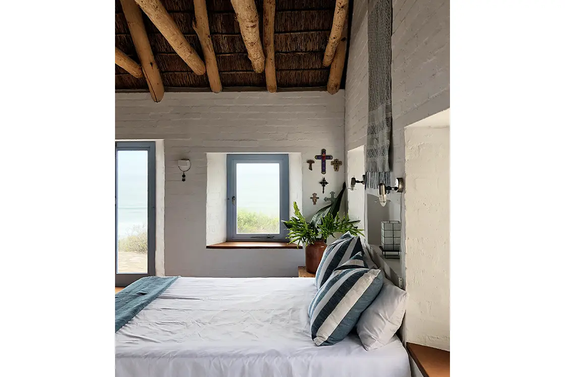 Perfect Hideaways property for sale: Cozy bedroom with rustic charm. Features a wooden beamed ceiling, white walls adorned with crosses, a window view of greenery, and a neatly made bed with striped pillows. Artemon Cottage, Paternoster.