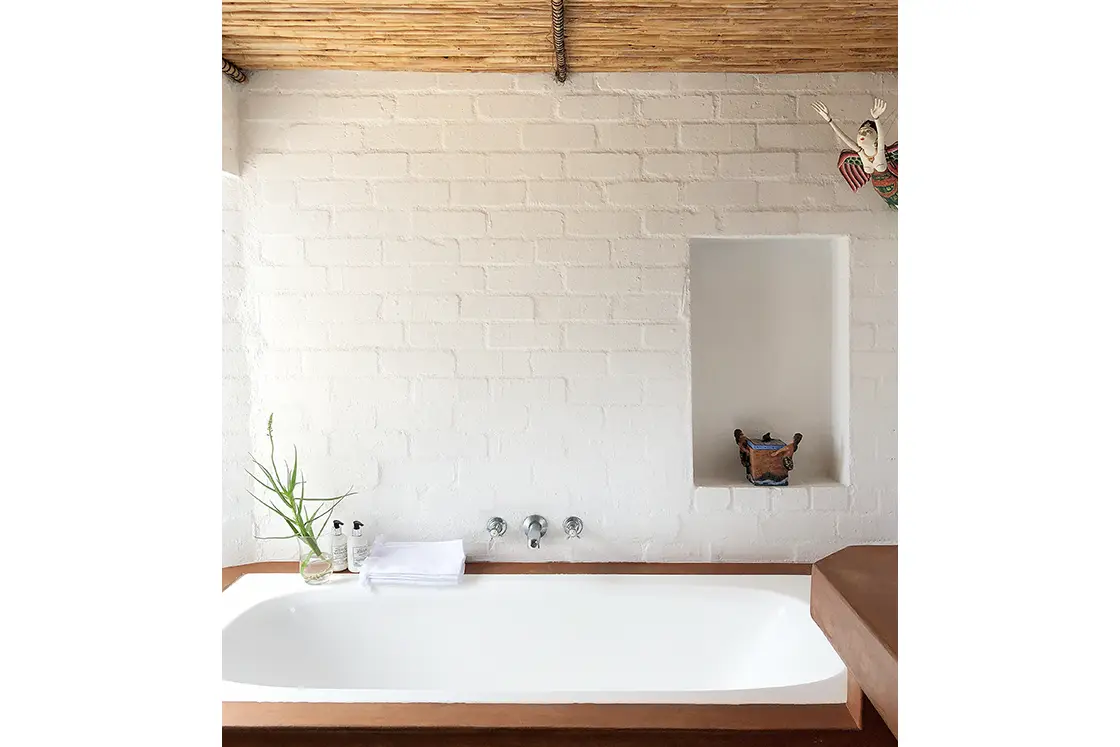 Perfect Hideaways property for sale: A minimalist bathroom with a white bathtub against a painted brick wall. A small niche holds a decorative item. Aloe plant, towels, and toiletries sit on the tub. Artemon Cottage, Paternoster.