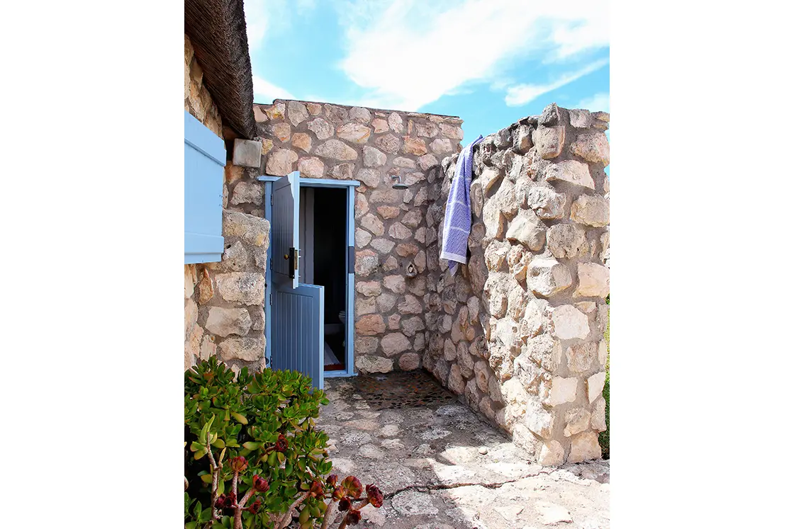 Perfect Hideaways property for sale: Stone cottage with blue door under a clear sky. A towel hangs on a wall, and greenery grows nearby. Artemon Cottage, Paternoster.