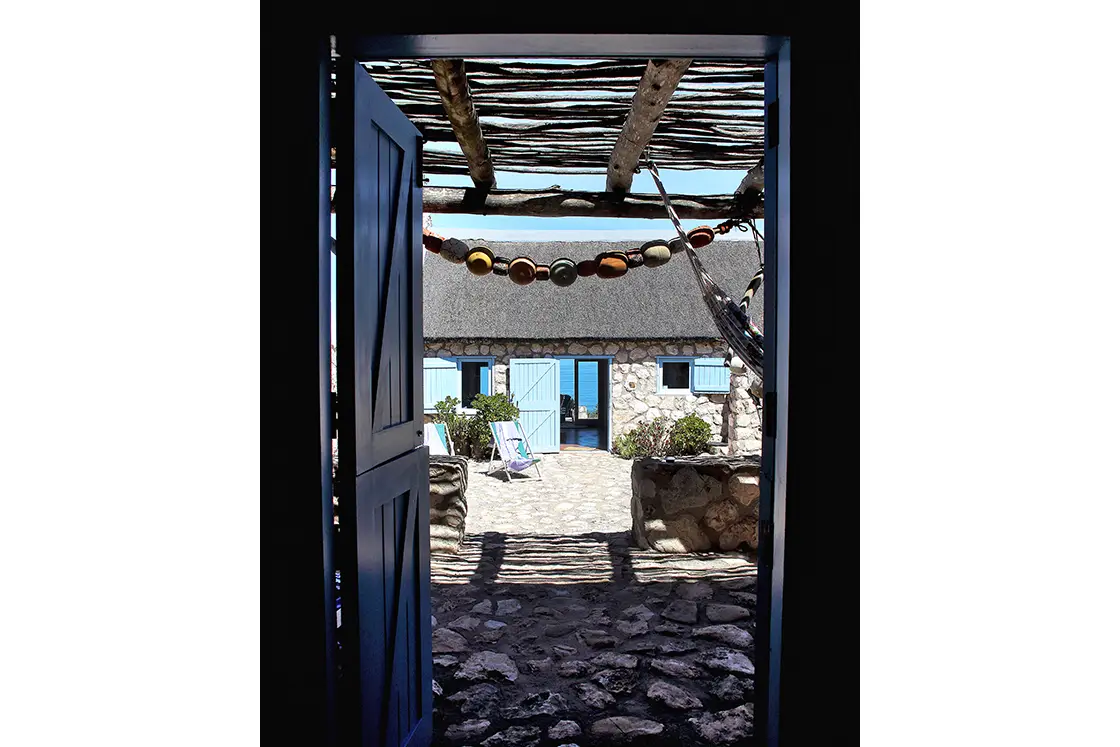 Perfect Hideaways property for sale: Peering through an open doorway, view of a sunlit stone courtyard with a thatched roof cottage. Blue shutters and loungers. Artemon Cottage, Paternoster.