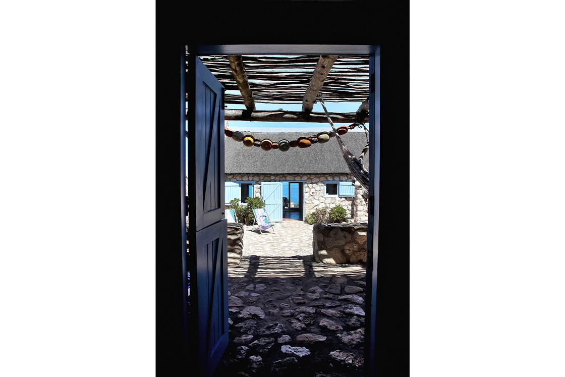 Perfect Hideaways property for sale: Peering through an open doorway, view of a sunlit stone courtyard with a thatched roof cottage. Blue shutters and loungers. Artemon Cottage, Paternoster.