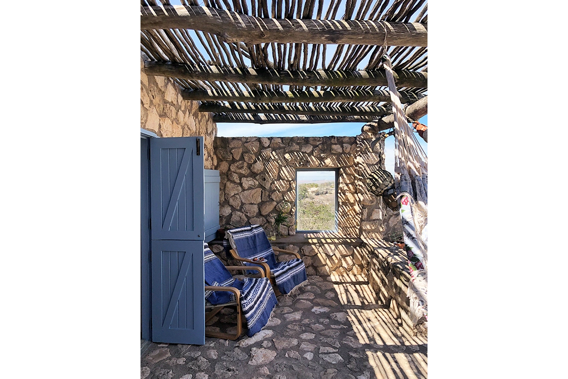 Perfect Hideaways property for sale: Rustic stone patio with woven wooden roof casting striped shadows. Blue cushioned chairs sit beside a window overlooking a desert landscape. Artemon Cottage, Paternoster.