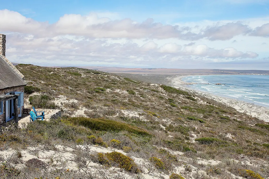 Perfect Hideaways property for sale: Coastal landscape with a stone cottage and blue chairs, overlooking a vast, sandy beach. The scene conveys tranquility under a bright blue sky. Artemon Cottage, Paternoster.