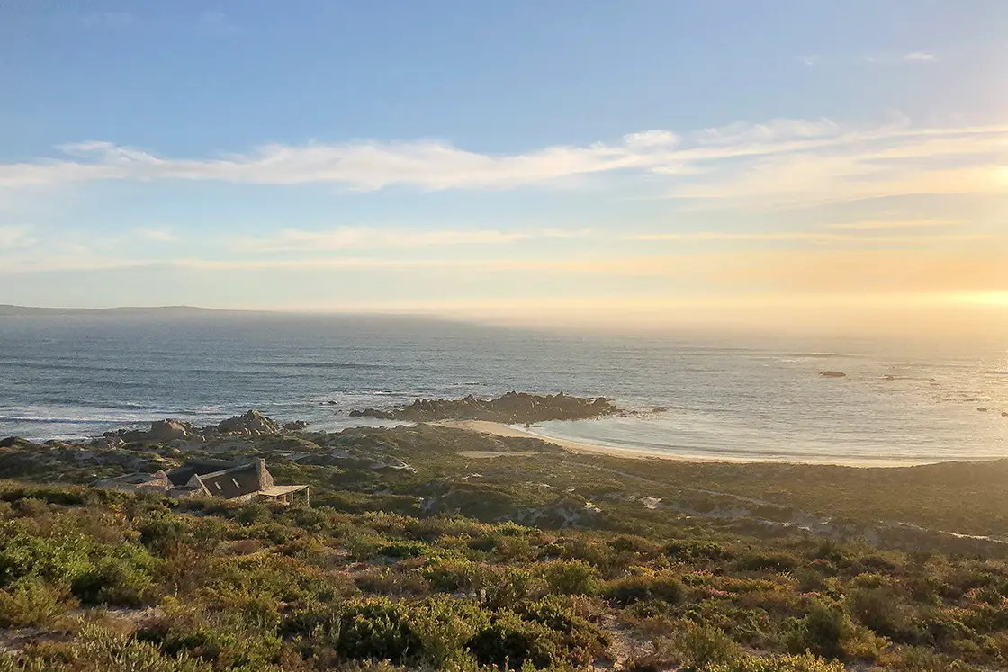 Perfect Hideaways property for sale: Coastal landscape at sunset, with a serene ocean meeting sandy shores. A house nestled in greenery. Soft sunlight. Artemon Cottage, Paternoster.