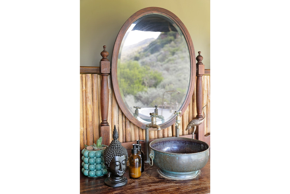 Perfect Hideaways real estate property: Rustic bathroom setup with a large oval mirror reflecting a scenic landscape. A bronze basin, antique faucet, Buddha sculpture, and soap pump are on a wooden counter, conveying a serene, natural vibe. Wolwehoek Private Nature Reserve, Montagu.