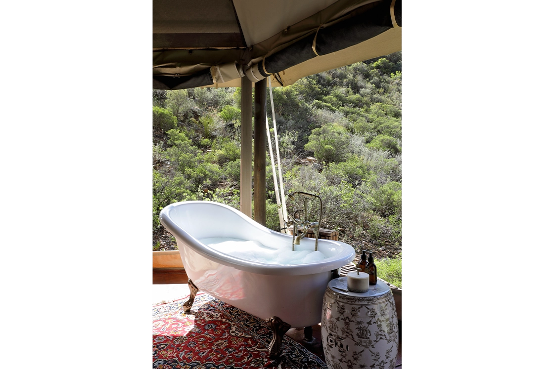 Perfect Hideaways real estate property: Outdoor bathtub with clawfoot design, filled with bubbles, overlooking lush greenery. Nearby, a small table holds candles and bottles, creating a serene, spa-like ambiance. Wolwehoek Private Nature Reserve, Montagu.