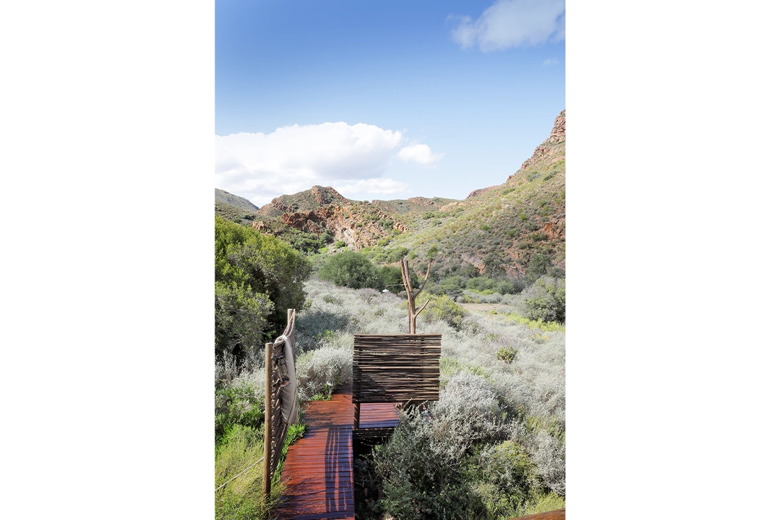 Perfect Hideaways real estate property: Remote wooden platform with rustic screens overlooks lush greenery and rugged, sunlit hills under a clear blue sky, conveying tranquility. Wolwehoek Private Nature Reserve, Montagu.