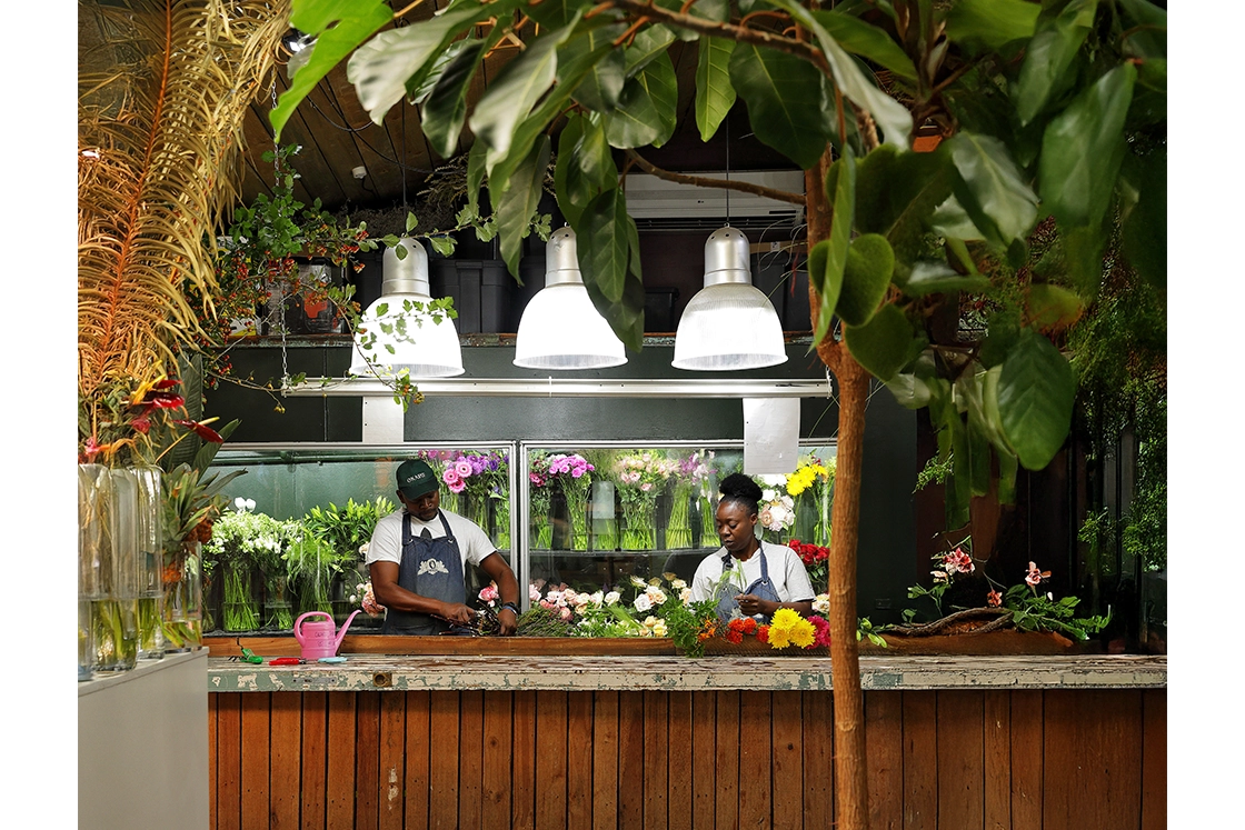 Perfect Hideaways property for sale: Two florists work in a lush flower shop, arranging colorful bouquets under bright hanging lights. The atmosphere is vibrant and serene. Okasie Florist, Stellenbosch.