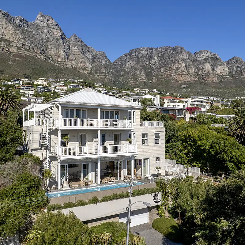 Perfect Hideaways property for sale: A luxurious three-story house with large balconies and a white facade is set against a backdrop of rugged mountains and surrounded by lush greenery. Sea Five, Camps Bay.