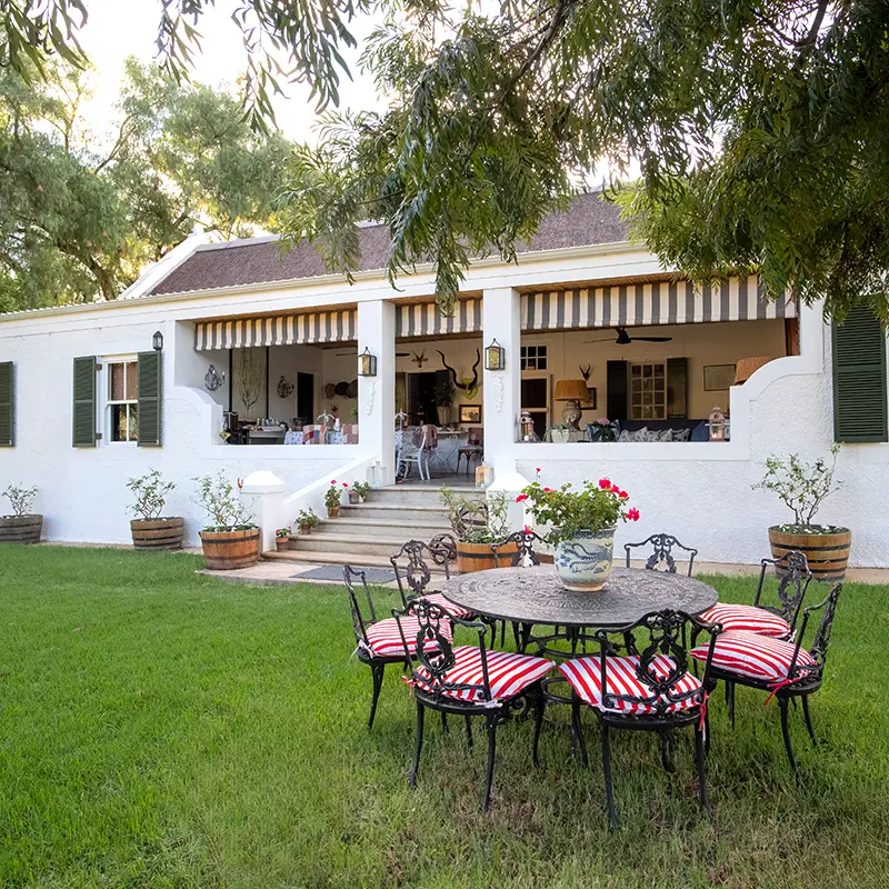 Daniel’s Kraal Farm. Perfect Hideaways property for sale: A charming white colonial-style house with a wide porch and green shutters. Red-cushioned iron chairs surround a patio table in the lush garden.