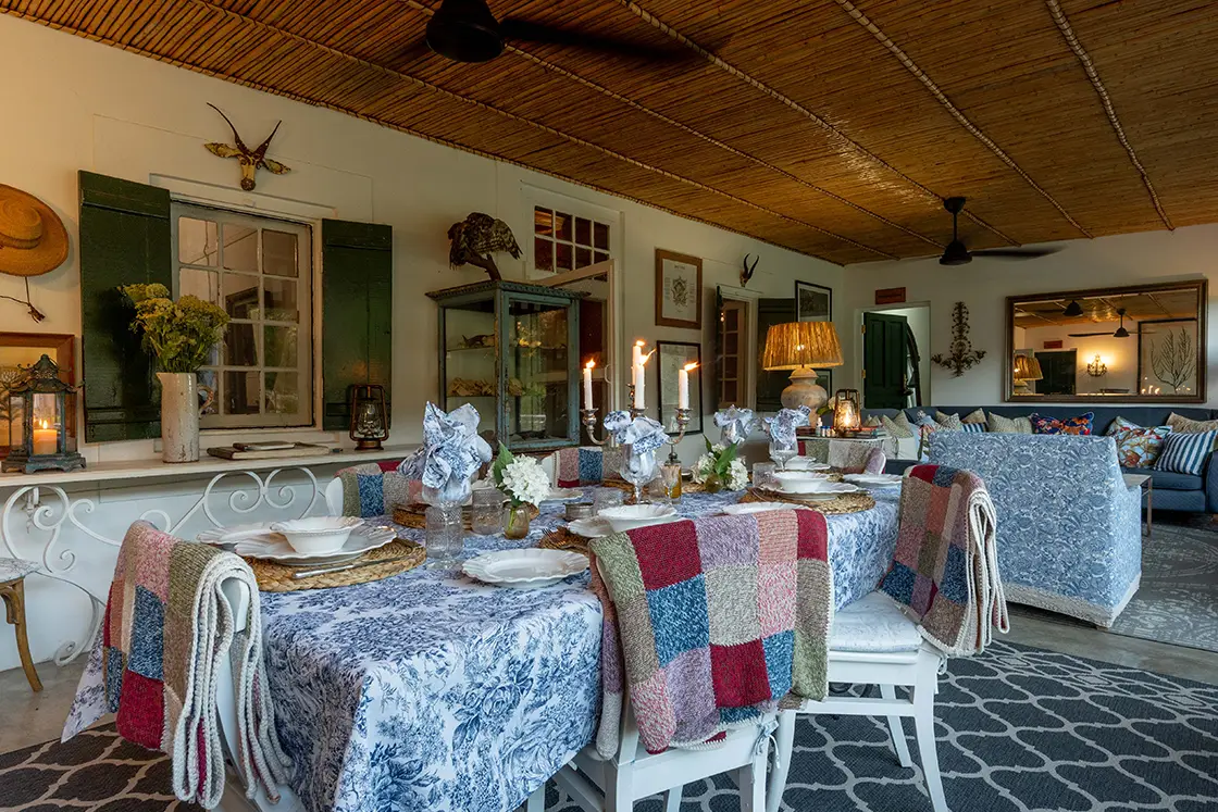 Perfect Hideaways property for sale: Elegant dining room with a rustic, cozy ambiance. A table with a blue floral tablecloth is set with candles, white plates, and colorful patchwork throws on chairs. Daniel’ Kraal Farm.